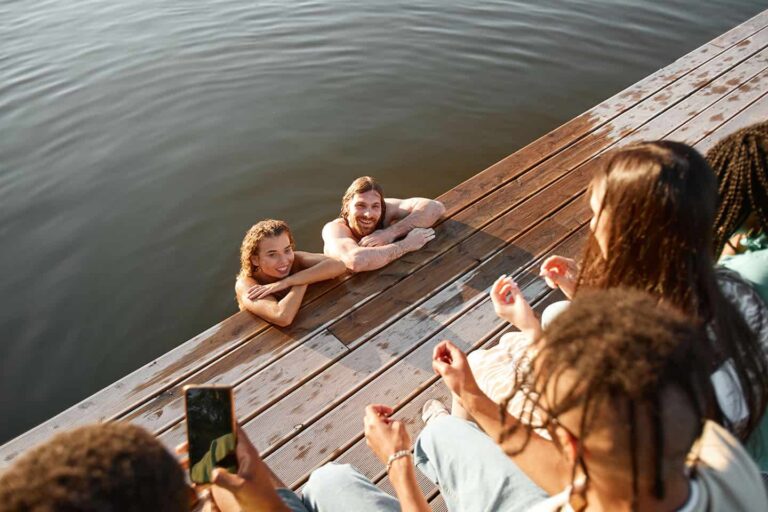 Group of friends gathered on a wooden dock while two people swim and smile at the camera