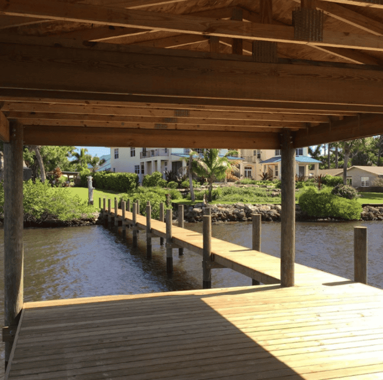 View from under a wooden boat house, looking out onto a long residential dock leading to a waterfront home surrounded by palm trees and landscaped greenery